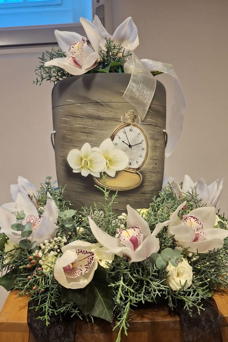 Eine graue Urne mit einer Uhr auf der Vorderseite eingebettet in einem Blumenkranz Eine graue Urne mit einer Uhr auf der Vorderseite eingebettet in einem Blumenkranz