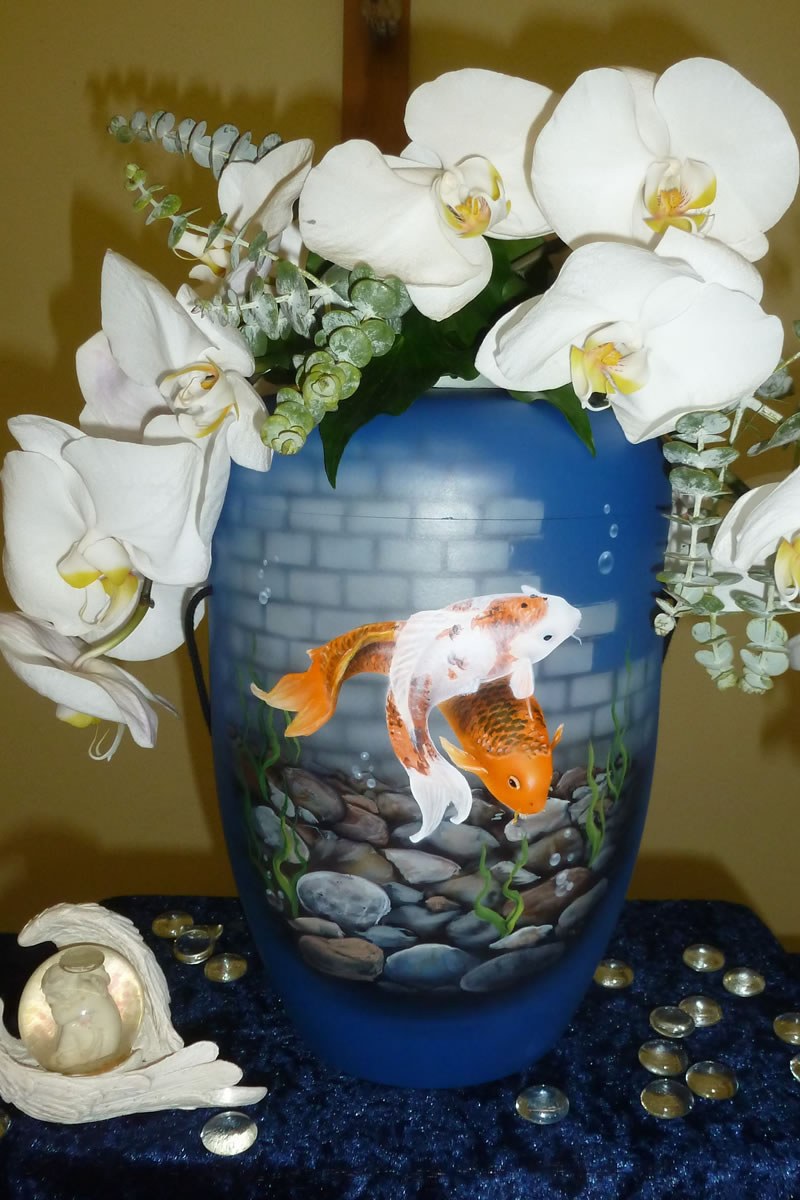 Eine hellblaue Urne mit zwei spielenden Fischen auf der Vorderseite umrahmt von weißen Blumen Eine hellblaue Urne mit zwei spielenden Fischen auf der Vorderseite umrahmt von weißen Blumen